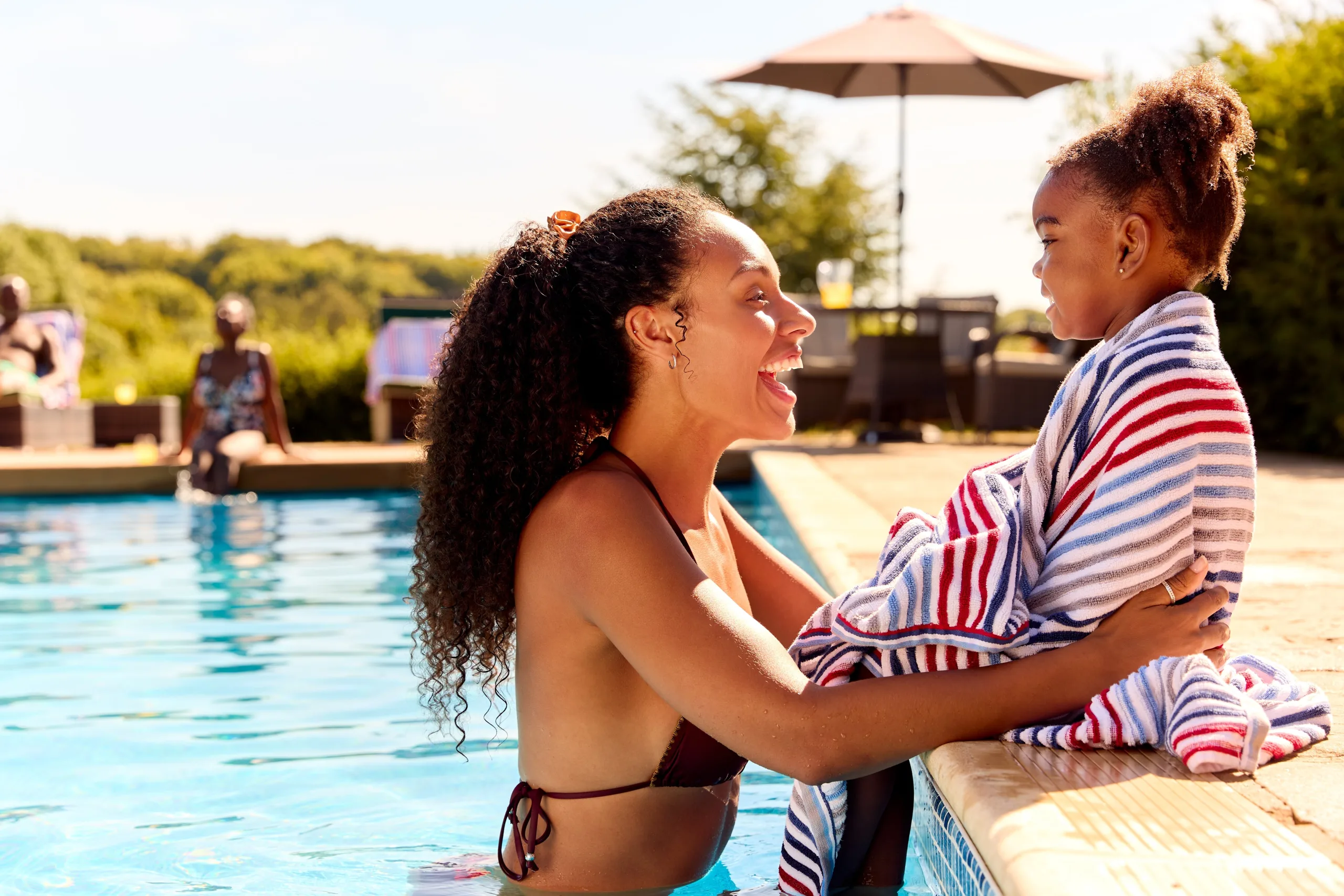 Mom and daughter laughing, mom in pool waist deep, daughter sitting on a ledge with toes in the pool wrapped in a towel looking at her mom. The sun is shining, and in the background, you can see a patio set with an umbrella and foliage.